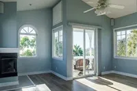 A bright room with blue-grey walls, a fireplace, hardwood floors, a ceiling fan, and large windows looking onto a balcony.