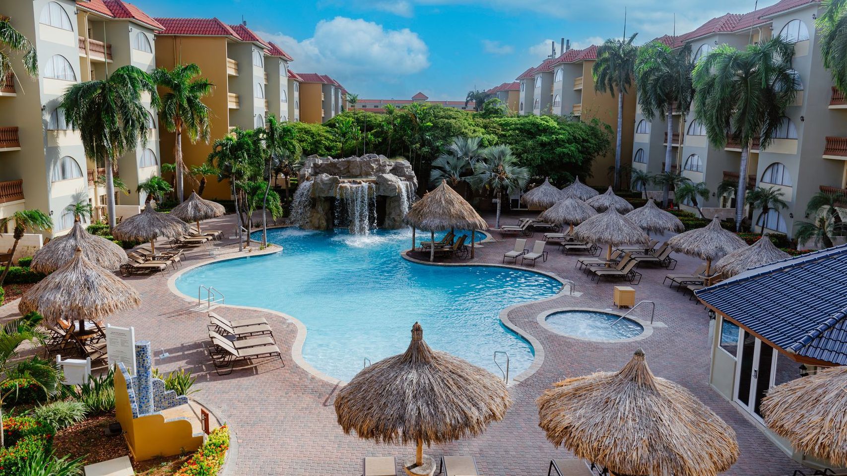 A large swimming pool surrounded by palm trees and umbrellas in a resort.