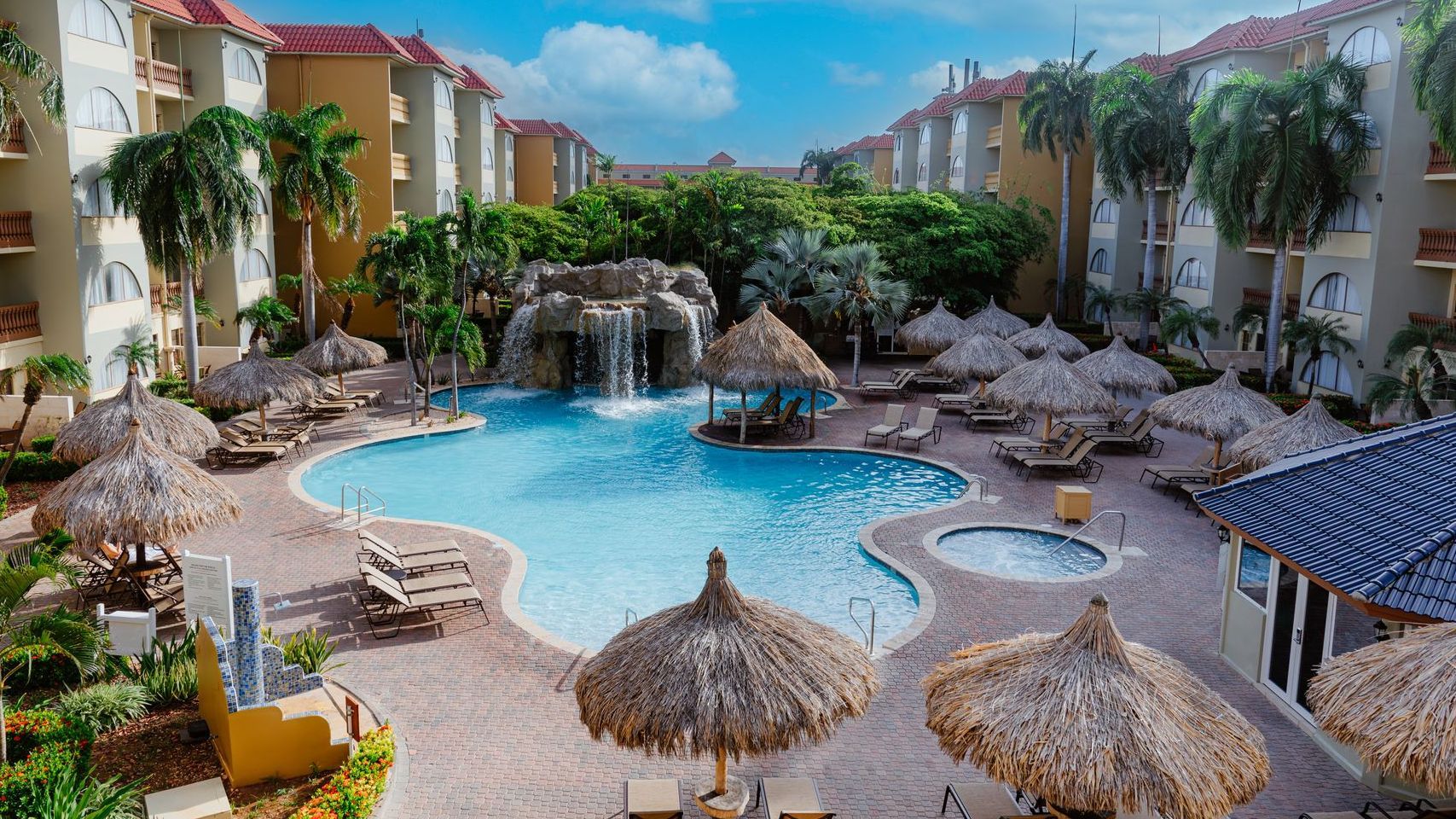 A large swimming pool surrounded by palm trees and umbrellas in a resort.