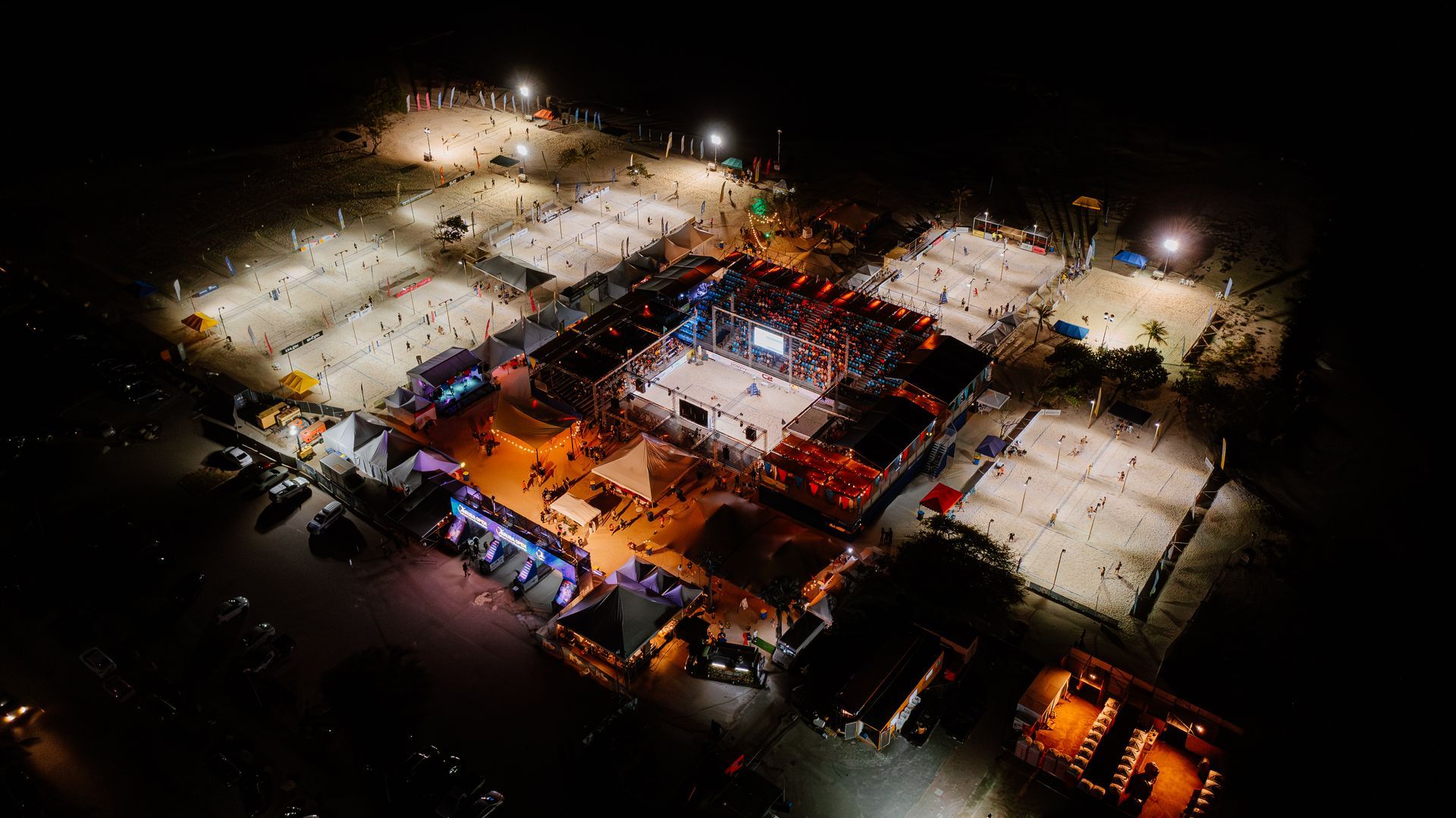 Nighttime aerial view of a lively beach volleyball event with lit courts, spectators, and structures.