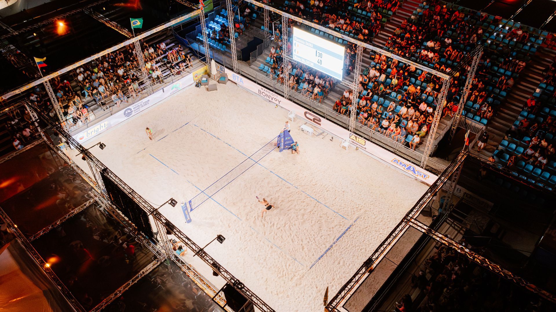 Aerial view of a beach volleyball court with players and spectators in an indoor stadium.