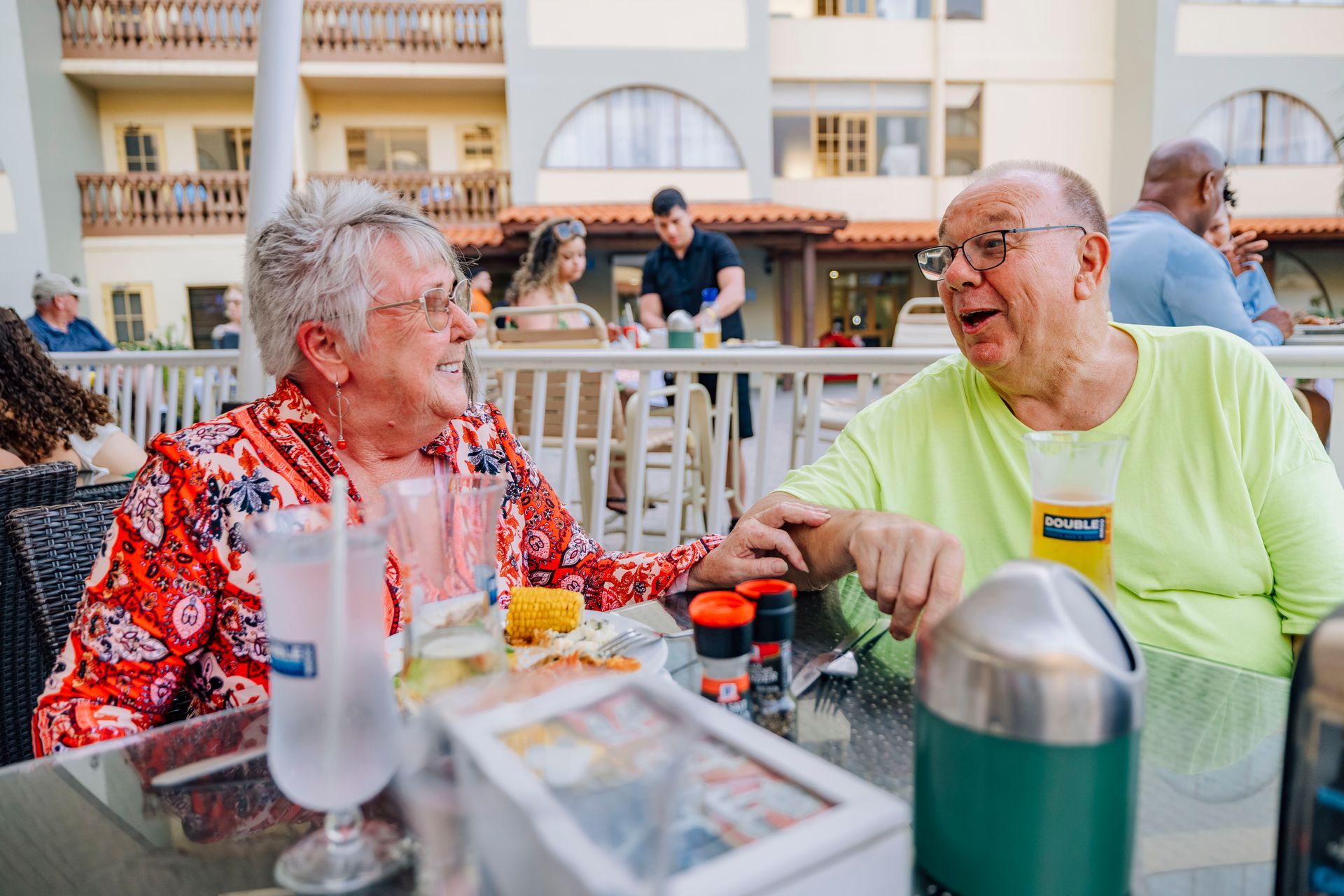 Elderly couple at an outdoor restaurant, smiling and conversing. The woman wears red and the man wears green.