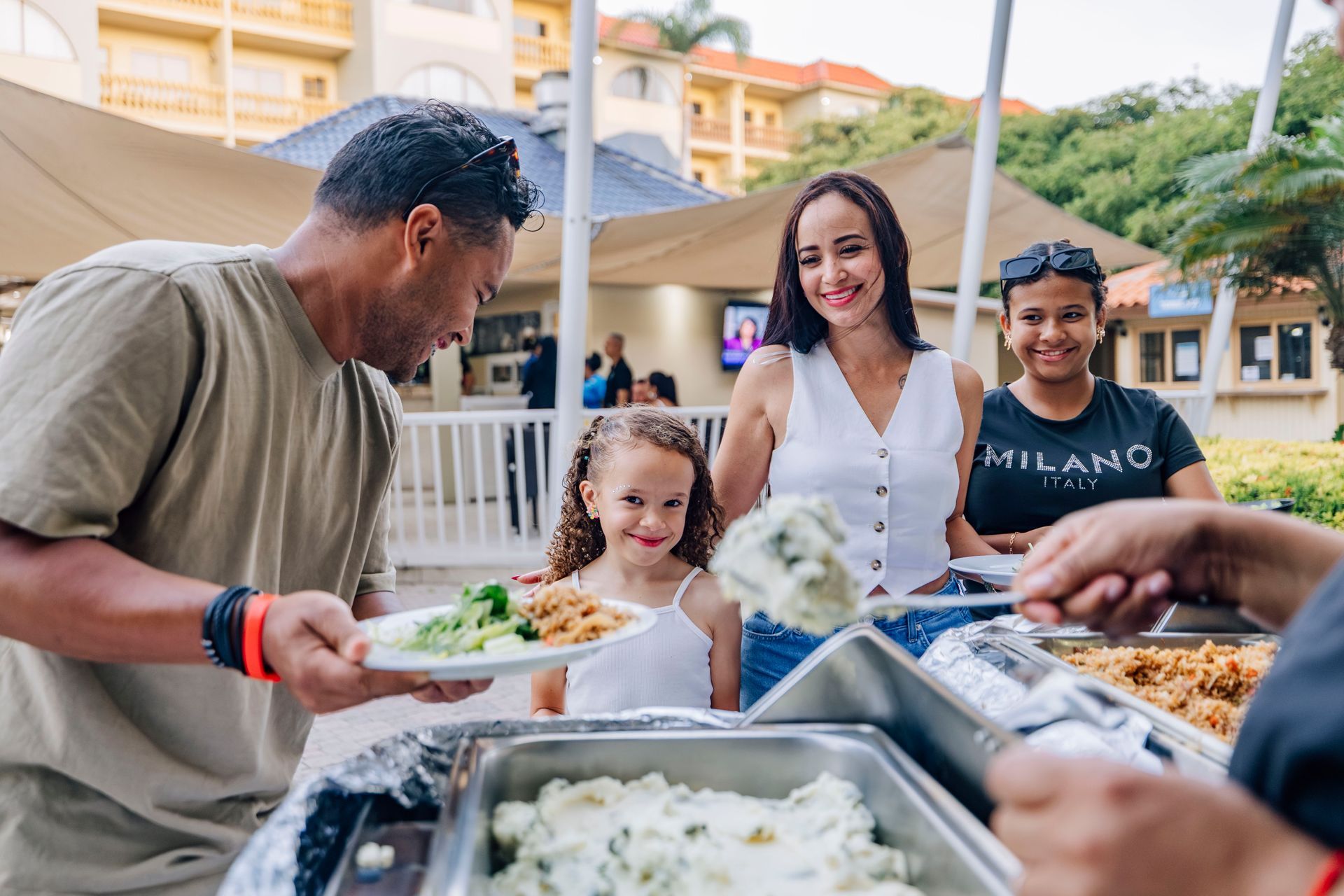 Family at a buffet, scooping food onto plates outdoors. Smiling and enjoying the meal.