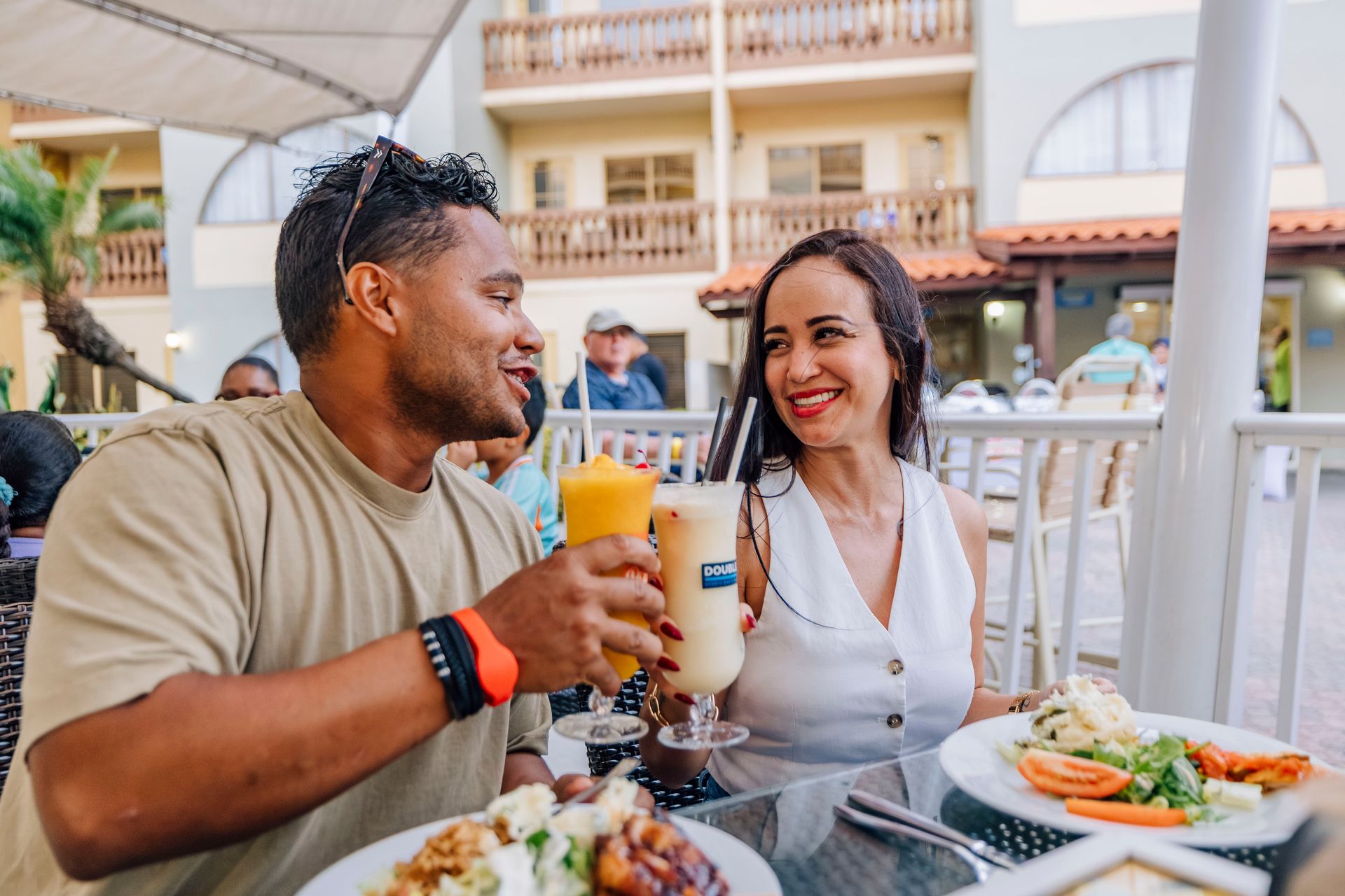 Couple toasting drinks at an outdoor restaurant, smiling. Food and tropical drinks visible.