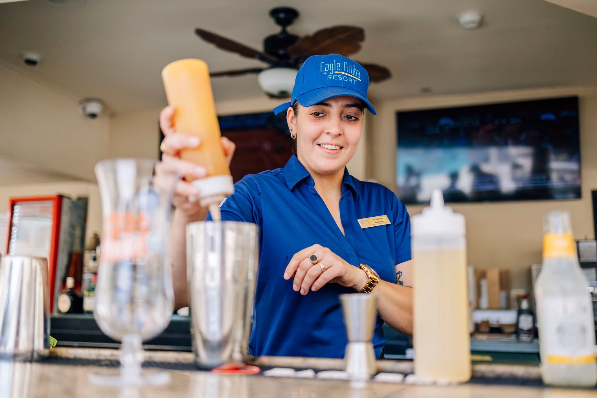 Bartender in blue uniform pouring liquid from a squeeze bottle into a shaker at an outdoor bar.