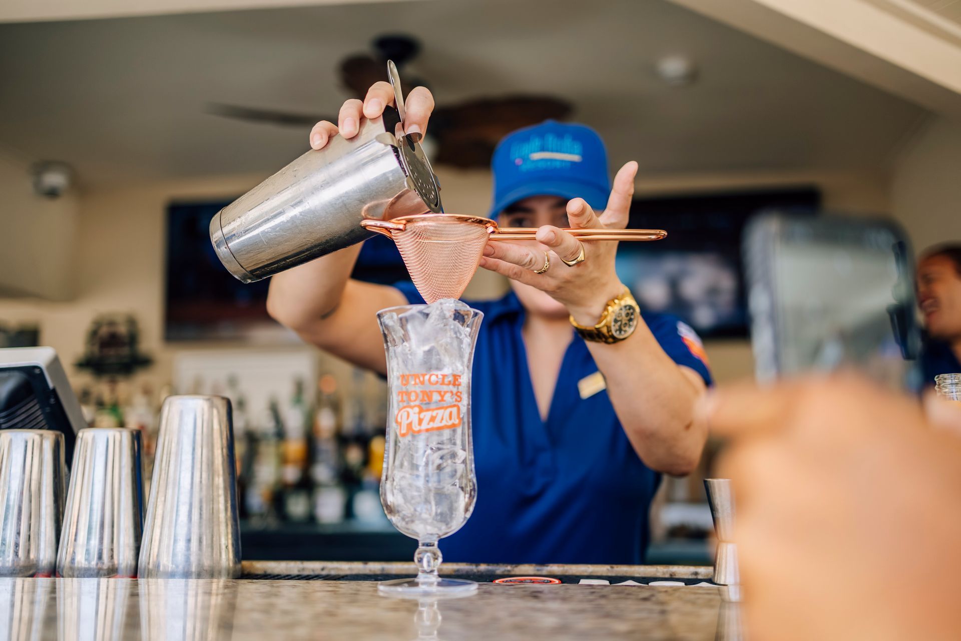 Bartender straining a pink cocktail into a tall glass, behind a bar with other bar tools.