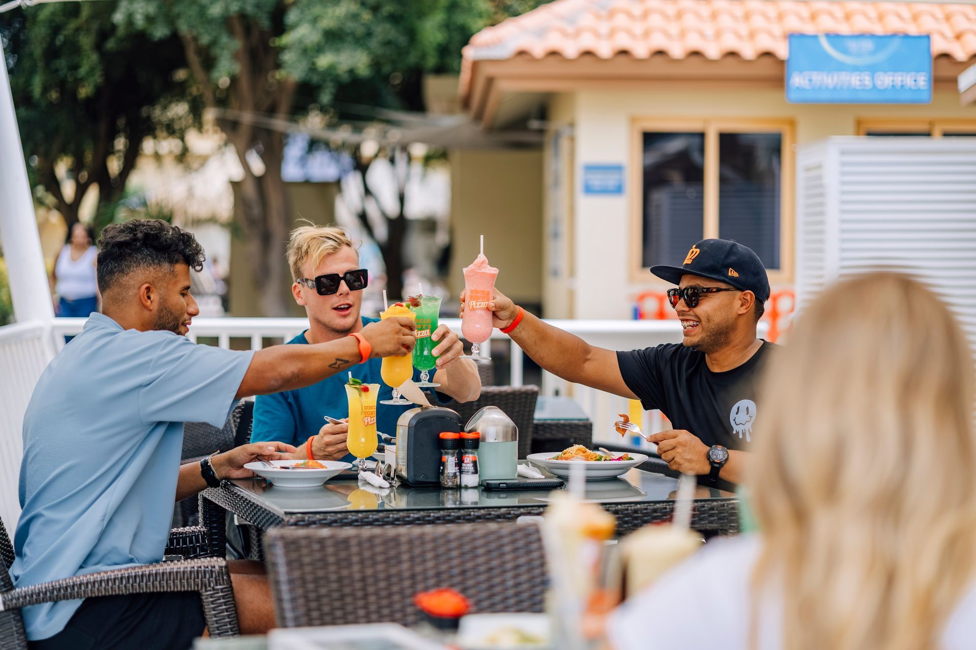 Four friends toasting drinks at an outdoor restaurant. One wears a black cap.