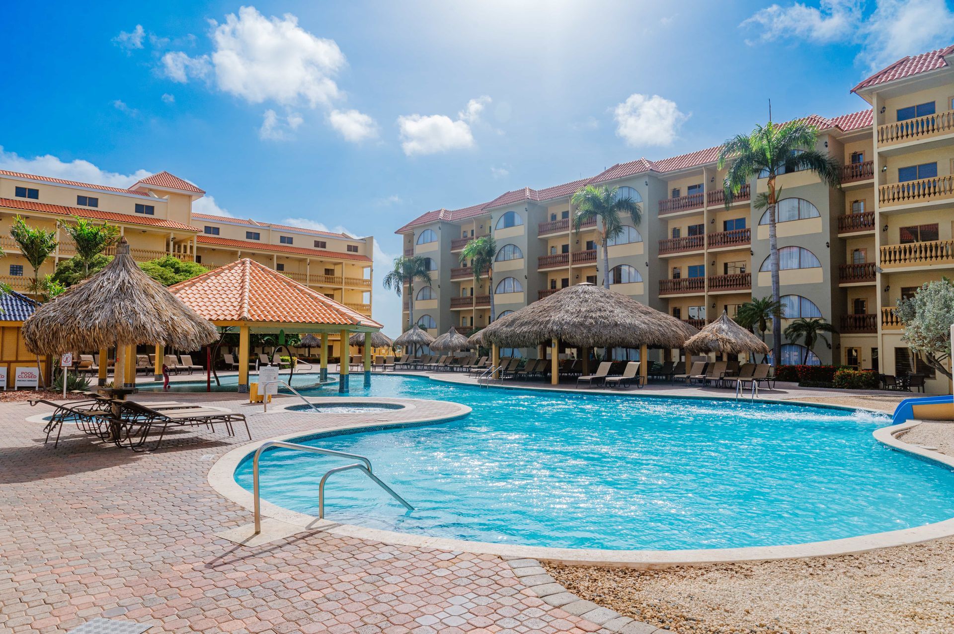 A large swimming pool in front of a hotel with thatched umbrellas.