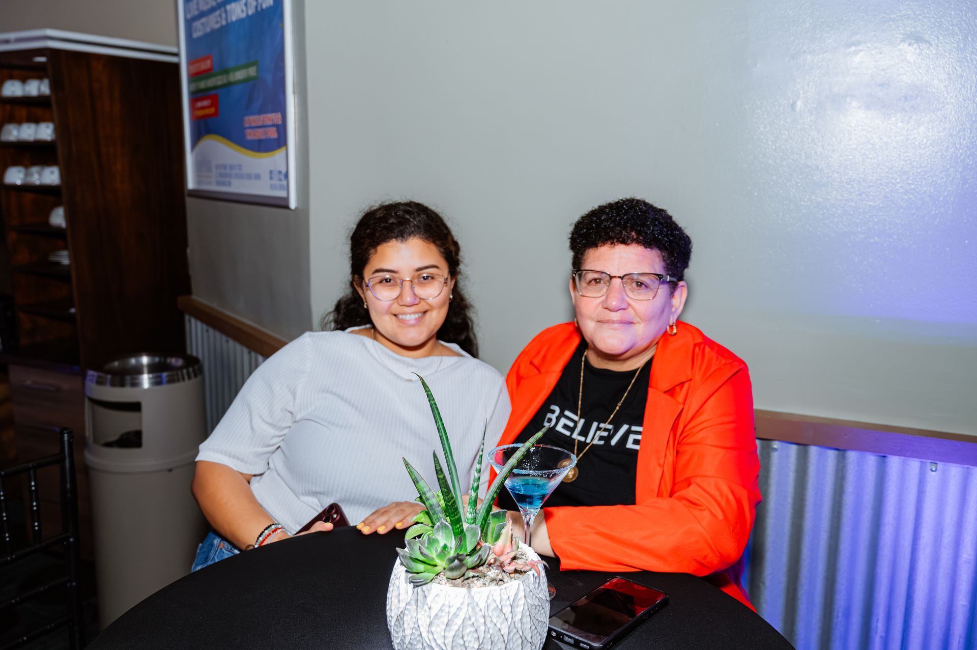 Two women are sitting at a table with a plant and a drink.