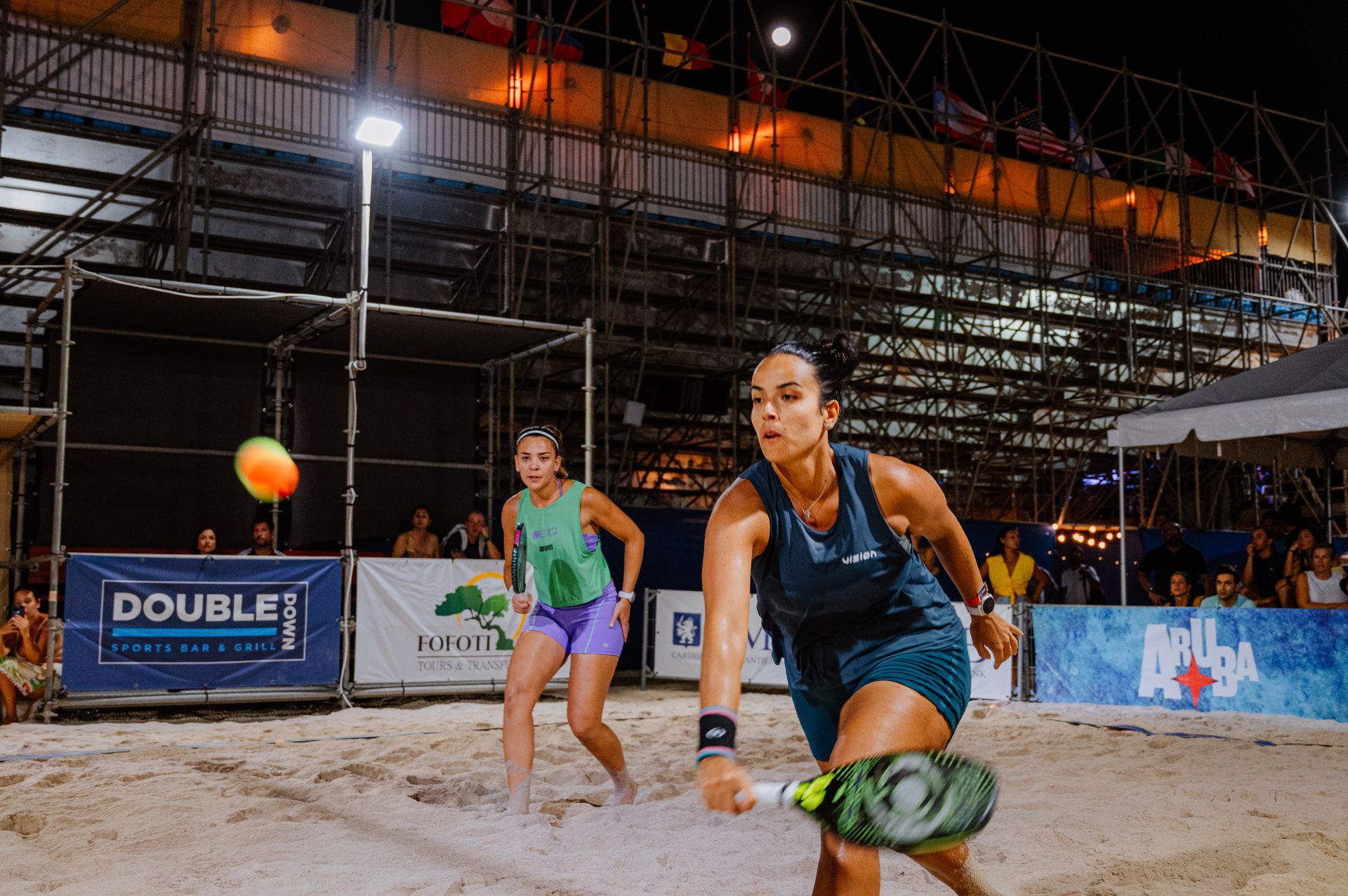 Woman in teal shirt hits beach tennis ball. Another woman watches in the background. Evening match.