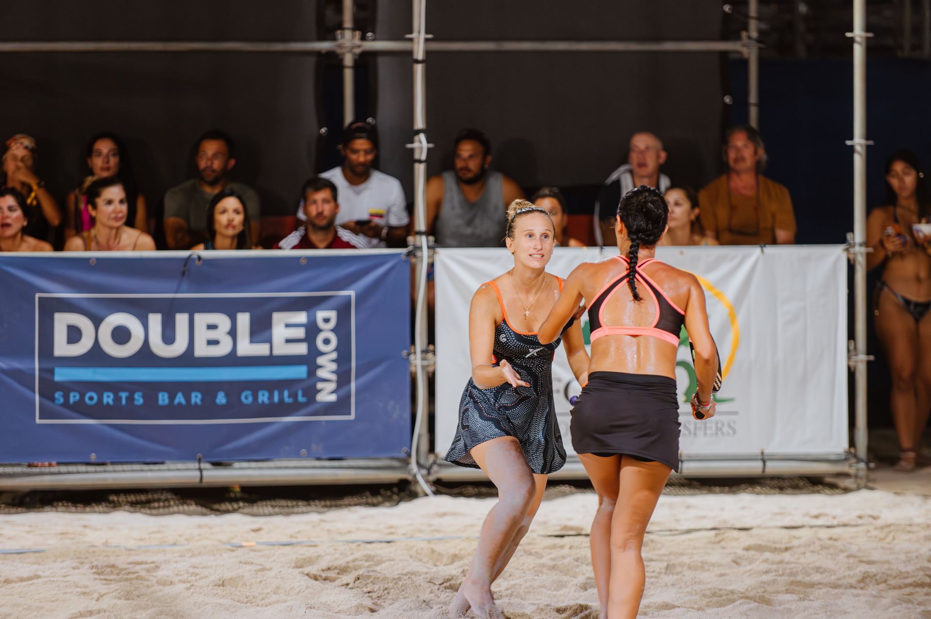 Two women in athletic wear on a sandy court; one reaches out, the other faces away. Spectators watch from above.
