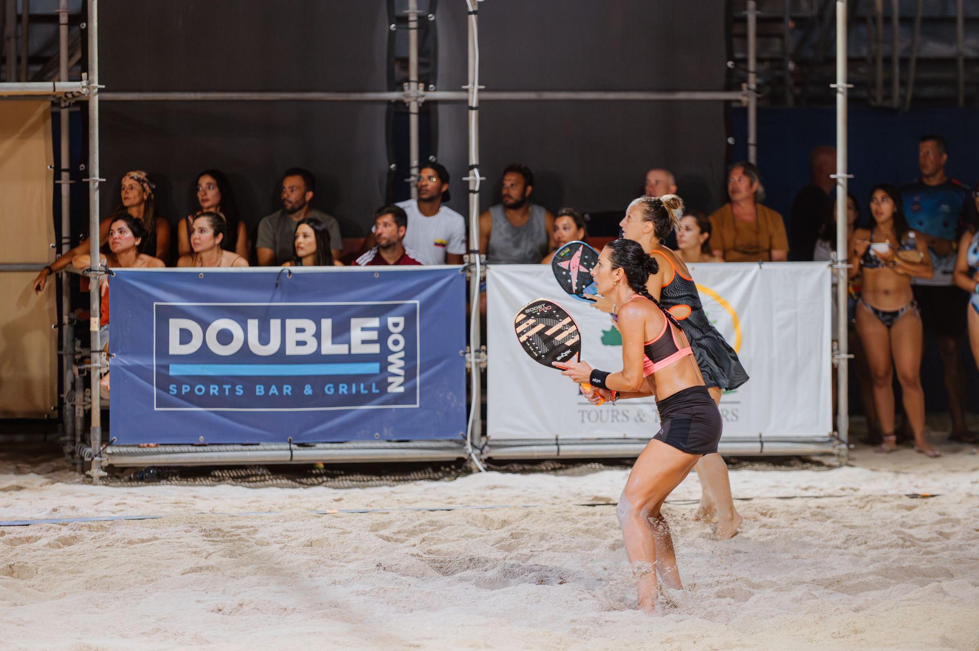 Two women playing beach tennis with spectators in a bar.