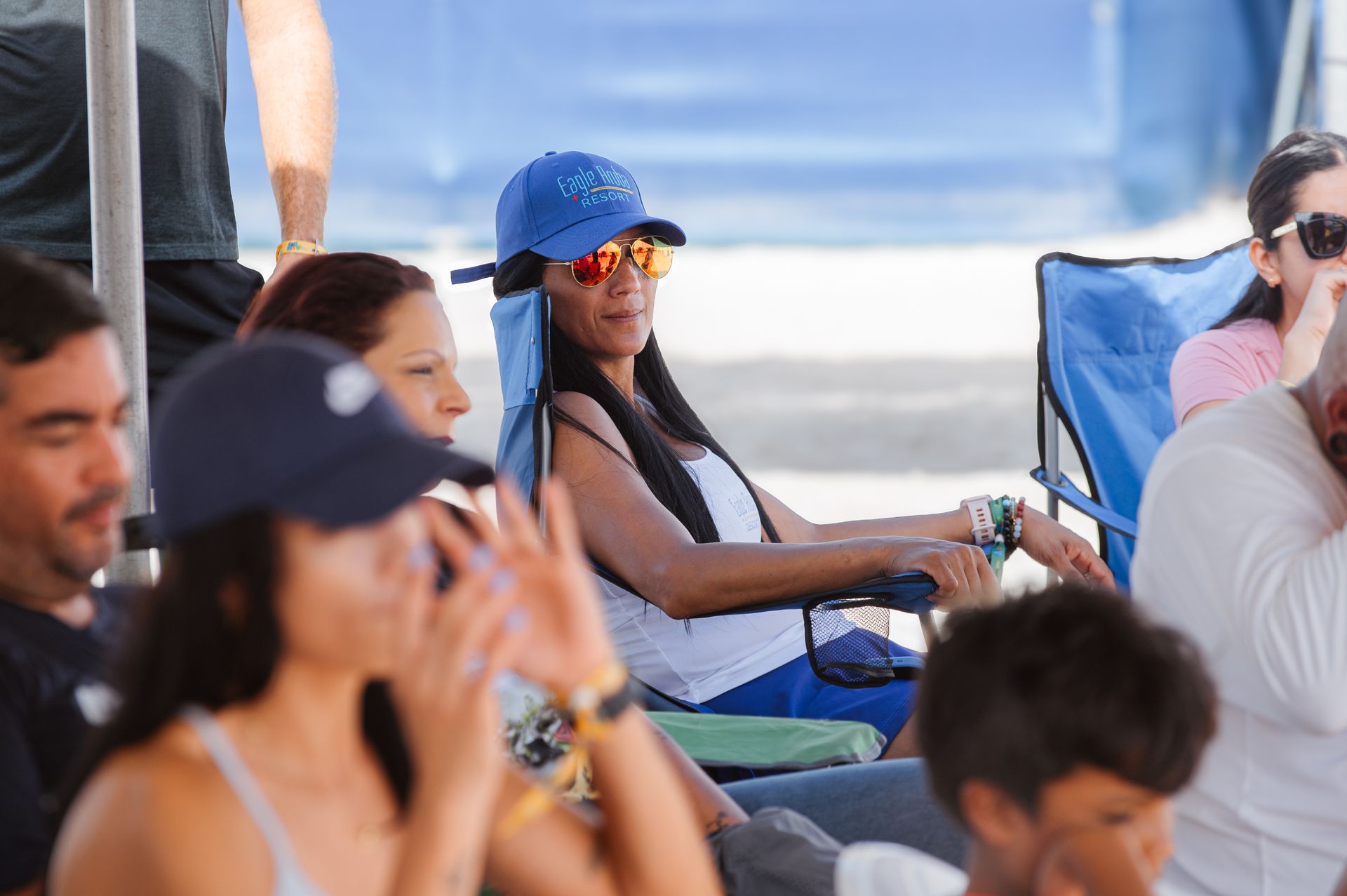 Woman in blue hat and sunglasses, sitting in a chair with other people outdoors.