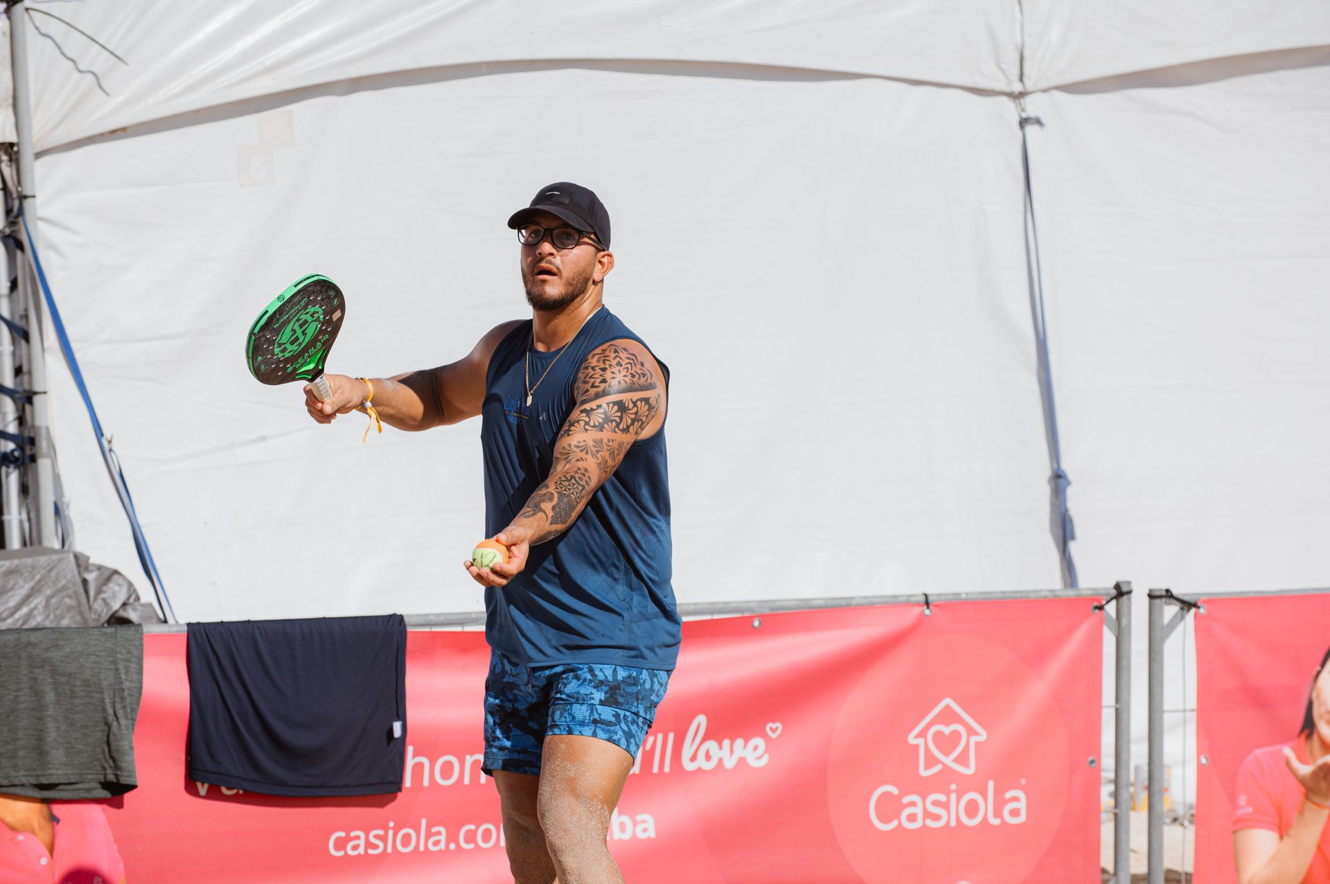 Man with arm tattoos playing beach tennis, holding a paddle and ball on a sandy court.