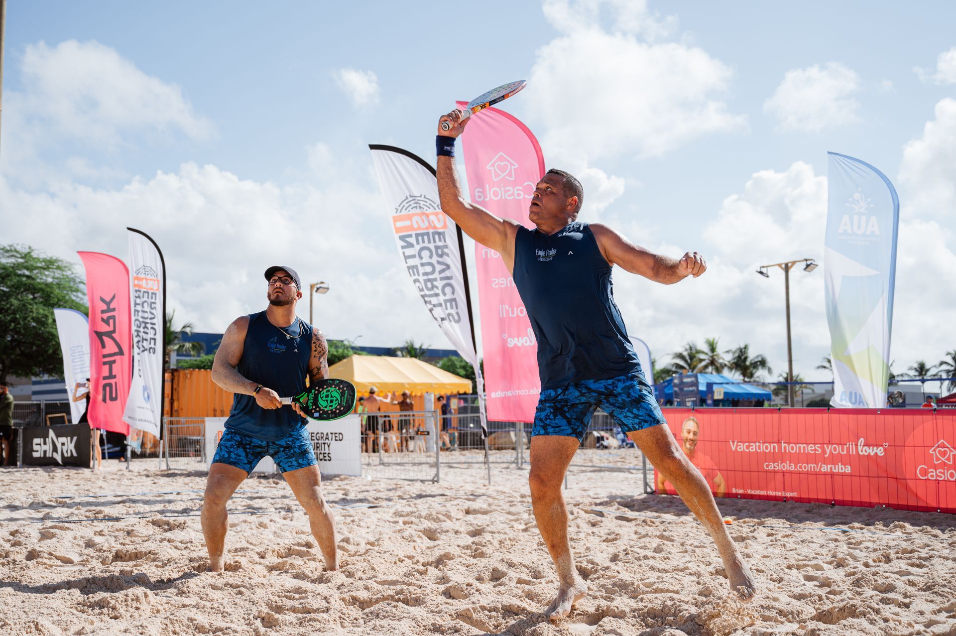 Two men playing beach tennis on a sunny day, one jumping and hitting a ball, the other ready.