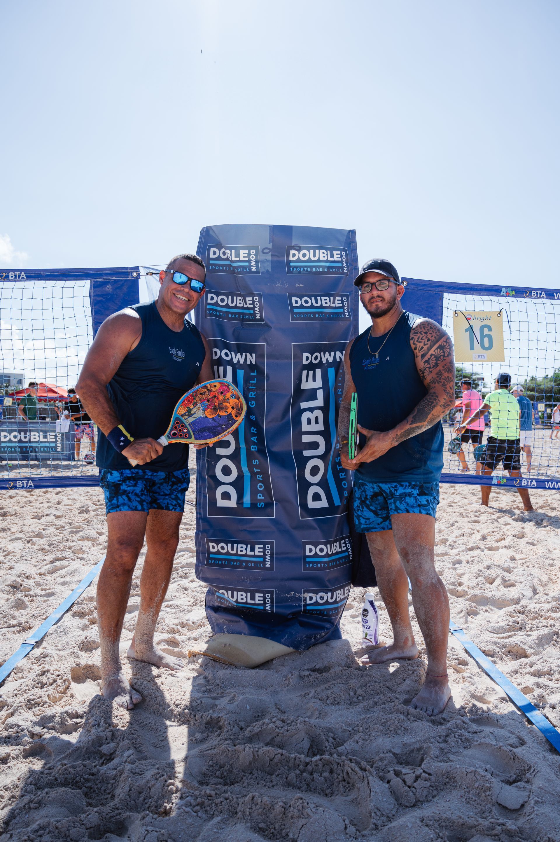 Two men on a beach holding a banner, one with a paddle. They wear blue shorts and tank tops on a sunny day.