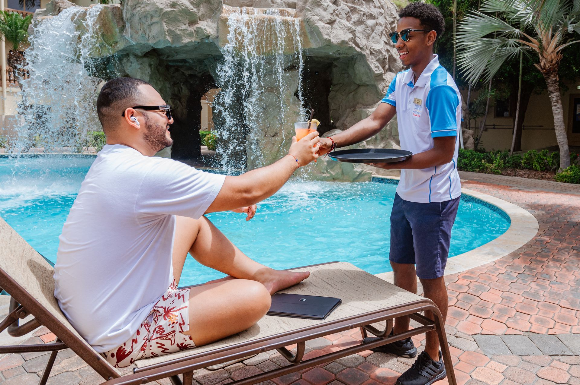 A man is sitting on a lounge chair next to a pool while a waiter serves him a drink.