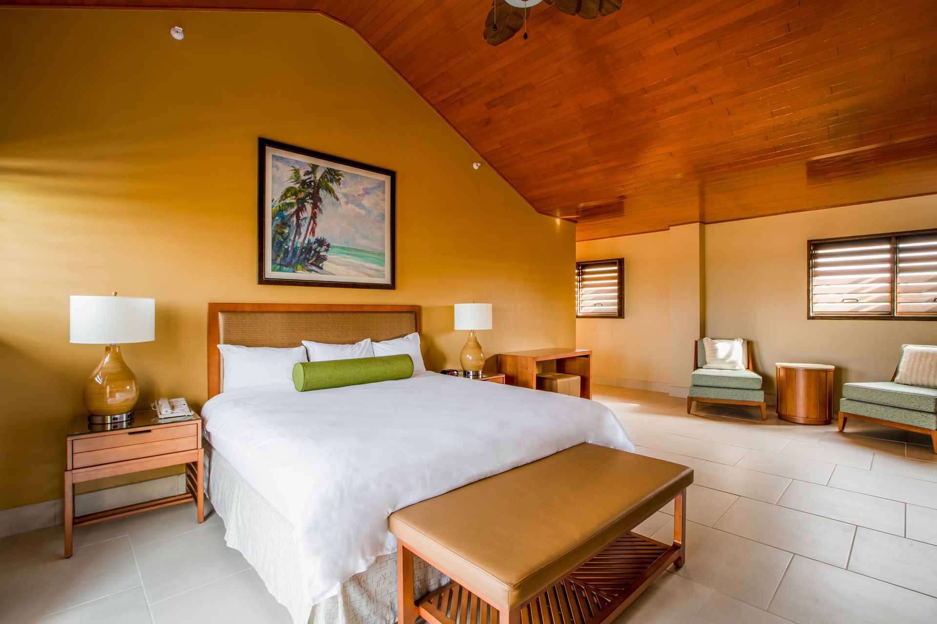 A hotel room with a king size bed , chairs , lamps and a bench.