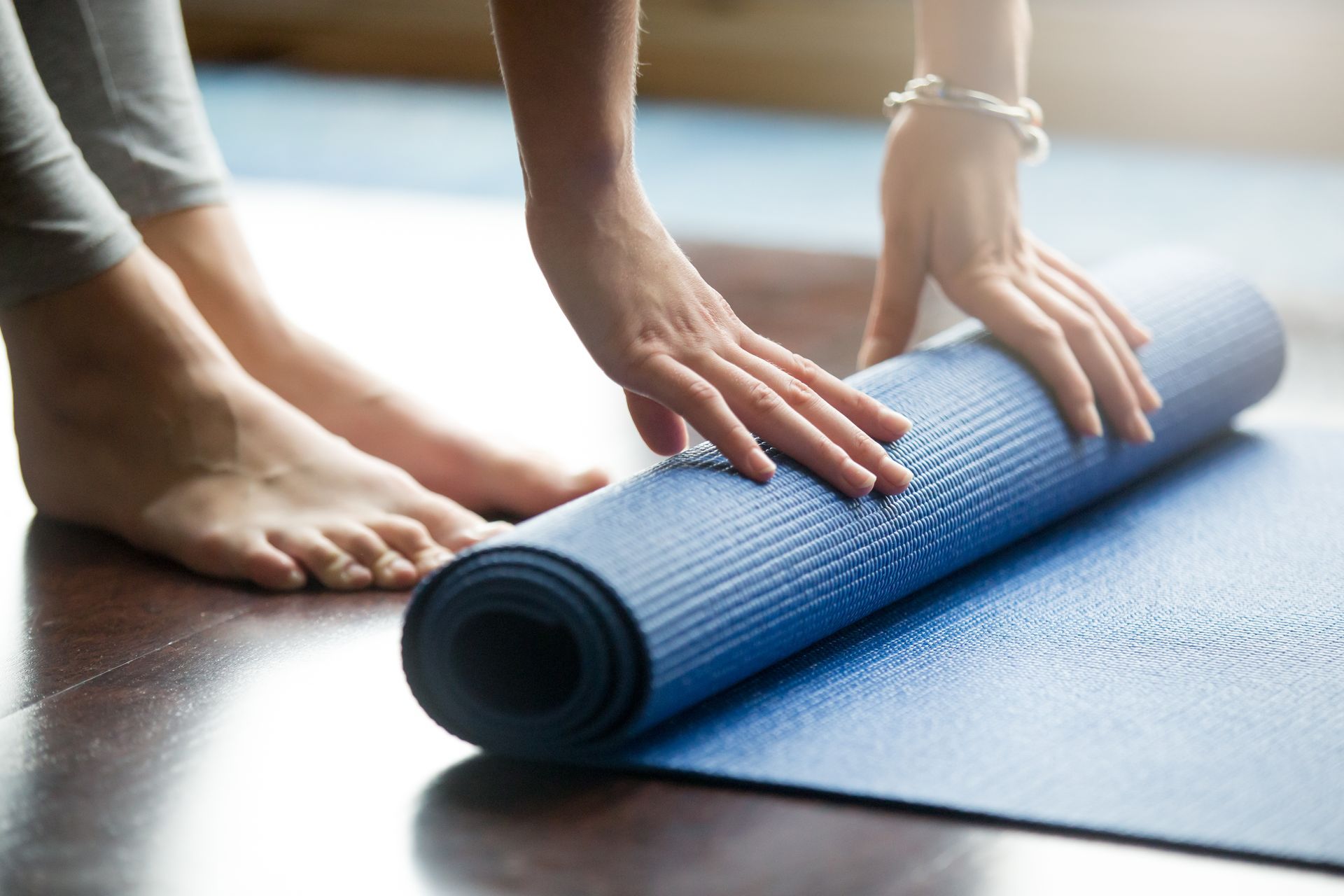 A woman is rolling a blue yoga mat on a wooden floor.