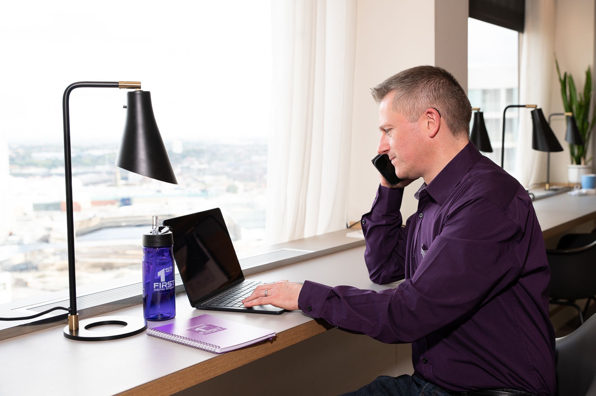 Andrew Wilson, Website Designer, is sitting at a desk using a laptop and talking on a cell phone.