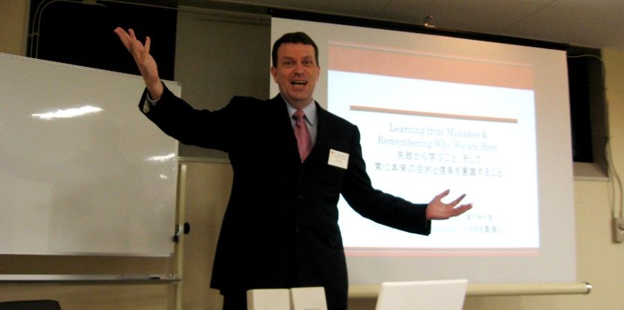 Ron Haigh giving a presentation with open arms. Slide with text and whiteboard are visible.