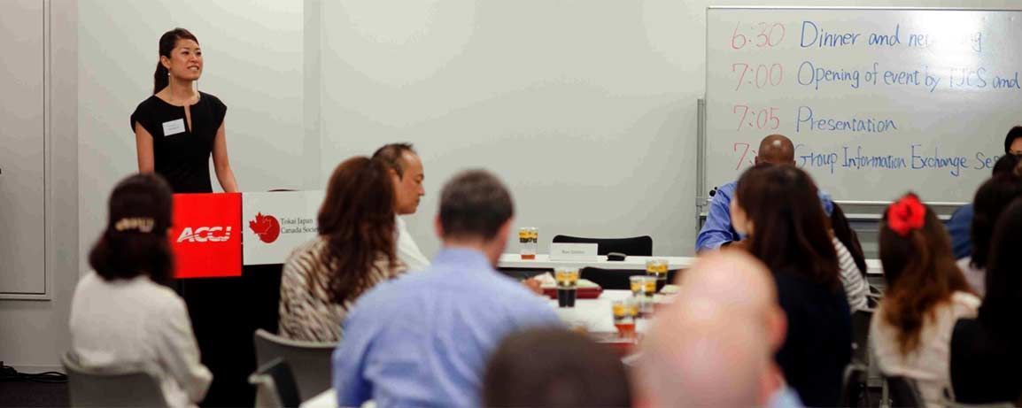 A woman giving a presentation, holding a red sign, at a meeting. Audience members are seated around tables.