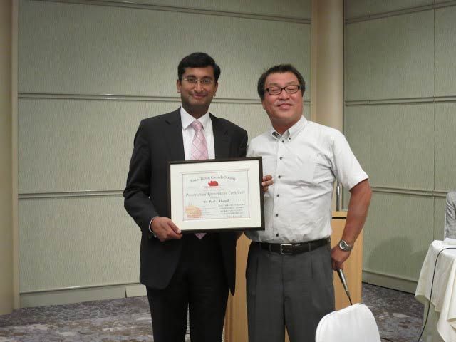 Two men, one in a suit, holding a framed certificate. The other man is wearing a polo shirt.