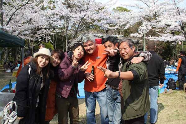 Group of people smiling, posing in front of cherry blossom trees. Outdoors on a sunny day.