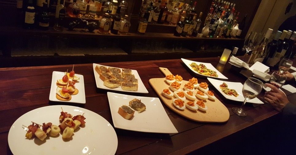 Appetizers on a bar counter with a liquor shelf backdrop. Various small dishes with colorful toppings.
