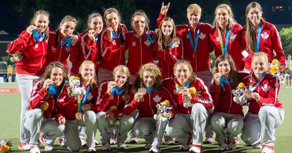 Canadian field hockey team celebrating with gold medals. Wearing red tracksuits, posing on a field.