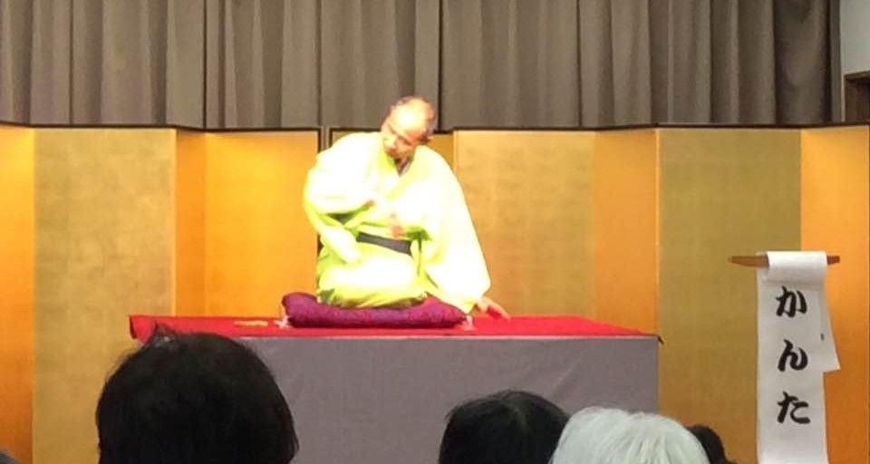 Man in yellow robe sitting on stage; gold backdrop, red table, and audience.