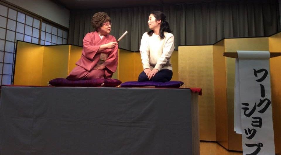 Two women on cushions, in front of gold screens. One holds a fan, and both appear to be conversing. 