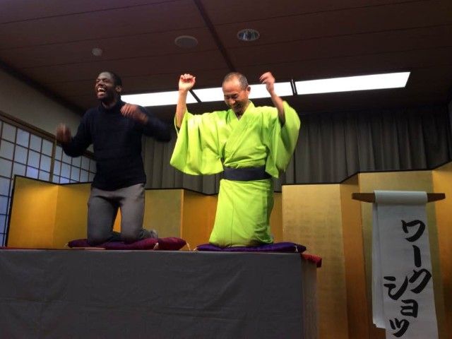 Two men kneeling, arms raised, on a stage with Japanese backdrop. One wears black, the other, a green robe.