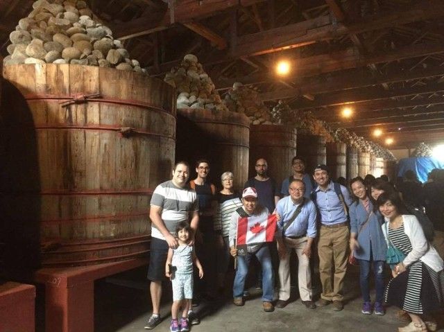 Group of people posing by large wooden fermentation vats, inside a rustic warehouse.