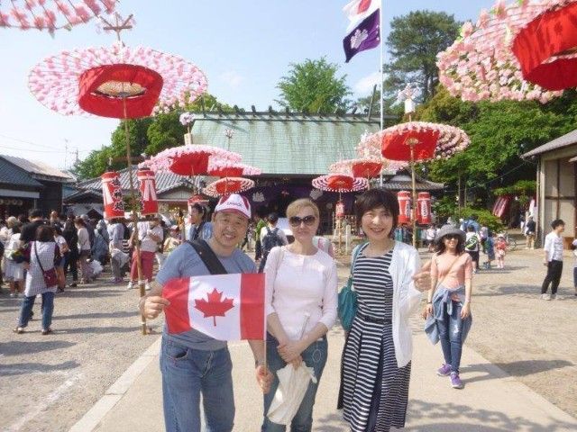 Three people holding a Canadian flag pose at a Japanese temple festival with red parasols and crowds.