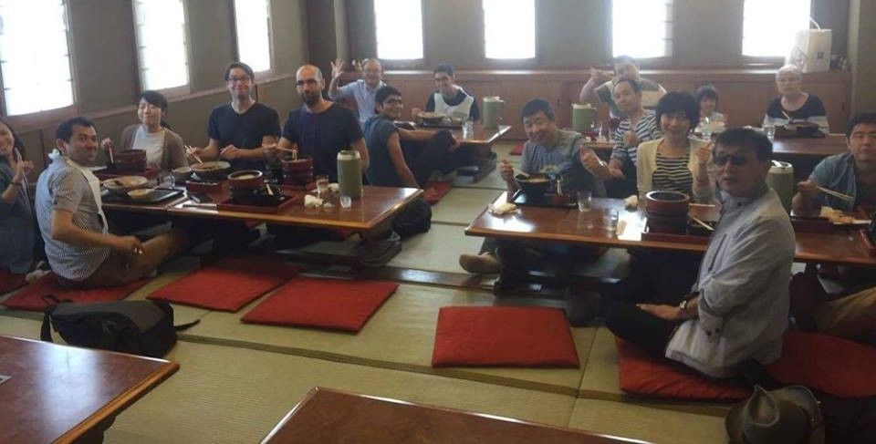 People sit at low tables in a Japanese restaurant. They eat, smile, and pose for a photo.