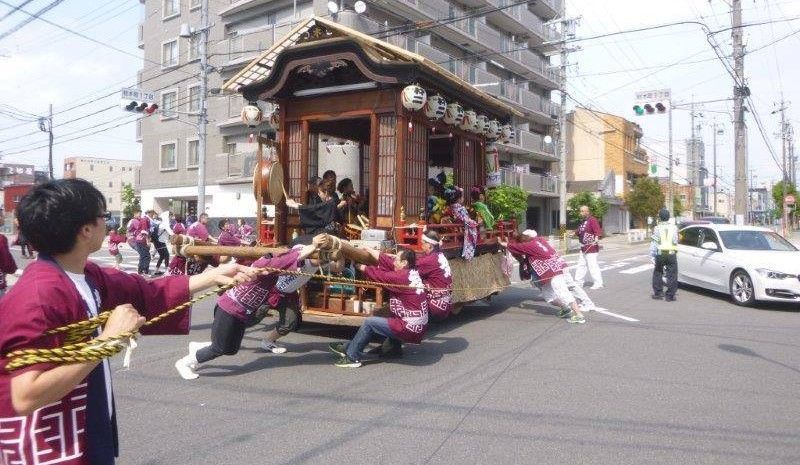 People pulling a large float with a wooden structure, in a city street during a festival.