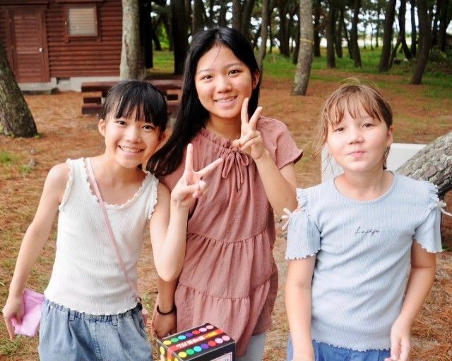 Three girls smiling in a park, center girl giving a peace sign.
