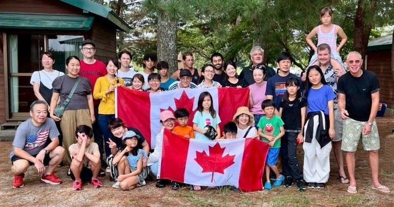 Group of people holding a Canadian flag posing outdoors near wooden cabins.