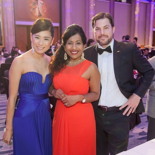 Three people pose together at a formal event. Woman in blue dress stands left, woman in red dress center, man in tuxedo right.