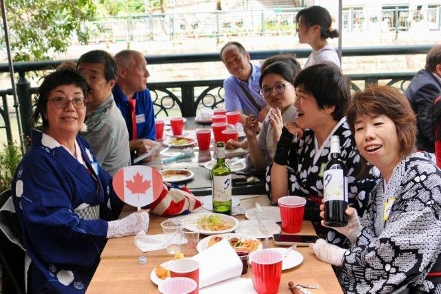 People seated at an outdoor table, some wearing kimonos, holding drinks and food, with a Canadian flag fan.