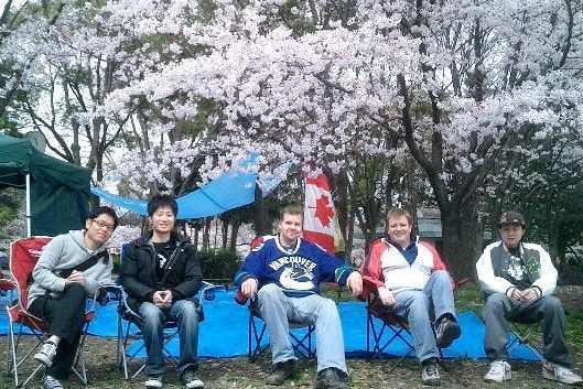 Five people seated in chairs under blooming cherry blossom trees with a Canadian flag.