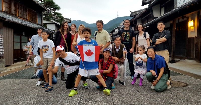 Group of people posing with a Canadian flag on a street in Japan, smiling. Wooden buildings in the background.