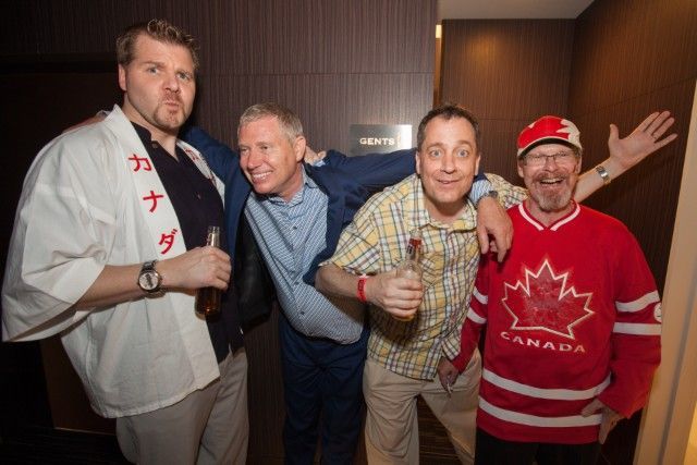 Four men smiling, posing together. One wears a Canada hockey jersey. Others hold drinks.