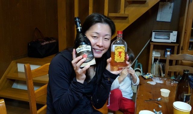 Woman holding a beer bottle and Fireball whiskey bottle, smiling indoors. Child visible nearby.
