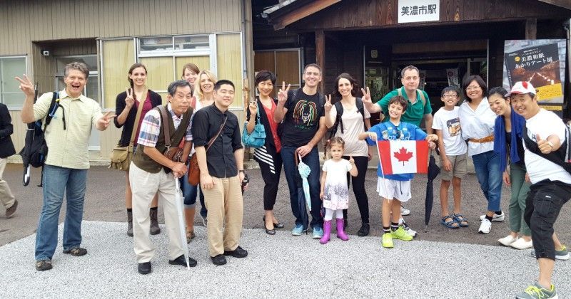 Group of people posing outdoors, some with peace signs. A small child holds a Canadian flag. Building in the background.