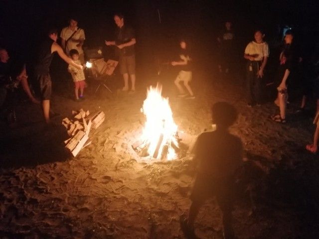 People gathered around a bonfire at night on a sandy beach, with chairs and a few standing nearby.