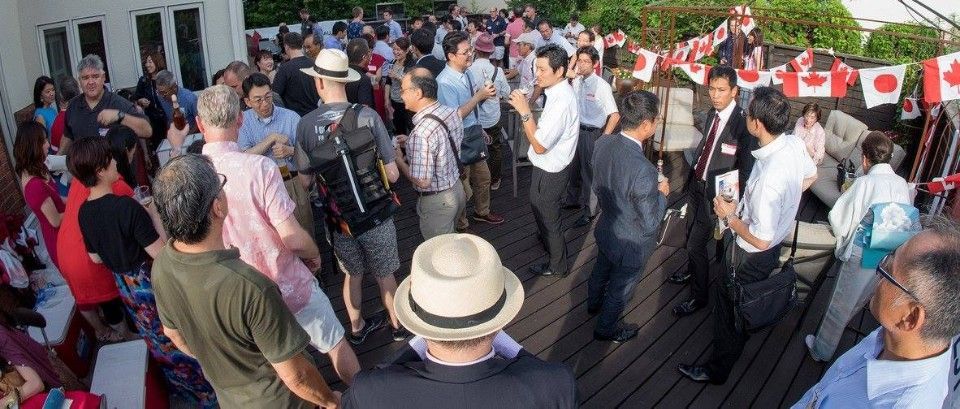 A crowded outdoor gathering with people socializing, wooden deck, flags, and a white building in the background.