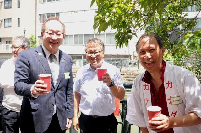 Three people smiling outdoors in front of a building, holding red cups in bright sunlight