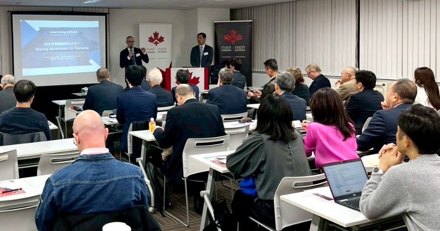 A presentation in a conference room. Two men speak at a podium, audience listens. Canadian flags are displayed.